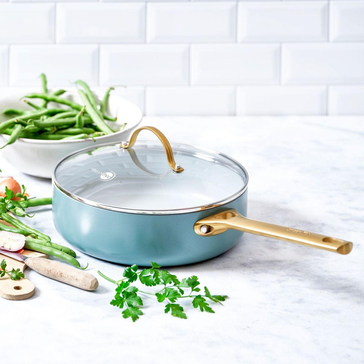 Eine blaue Pfanne mit goldenem Griff und Glasdeckel, daneben grüne Bohnen.