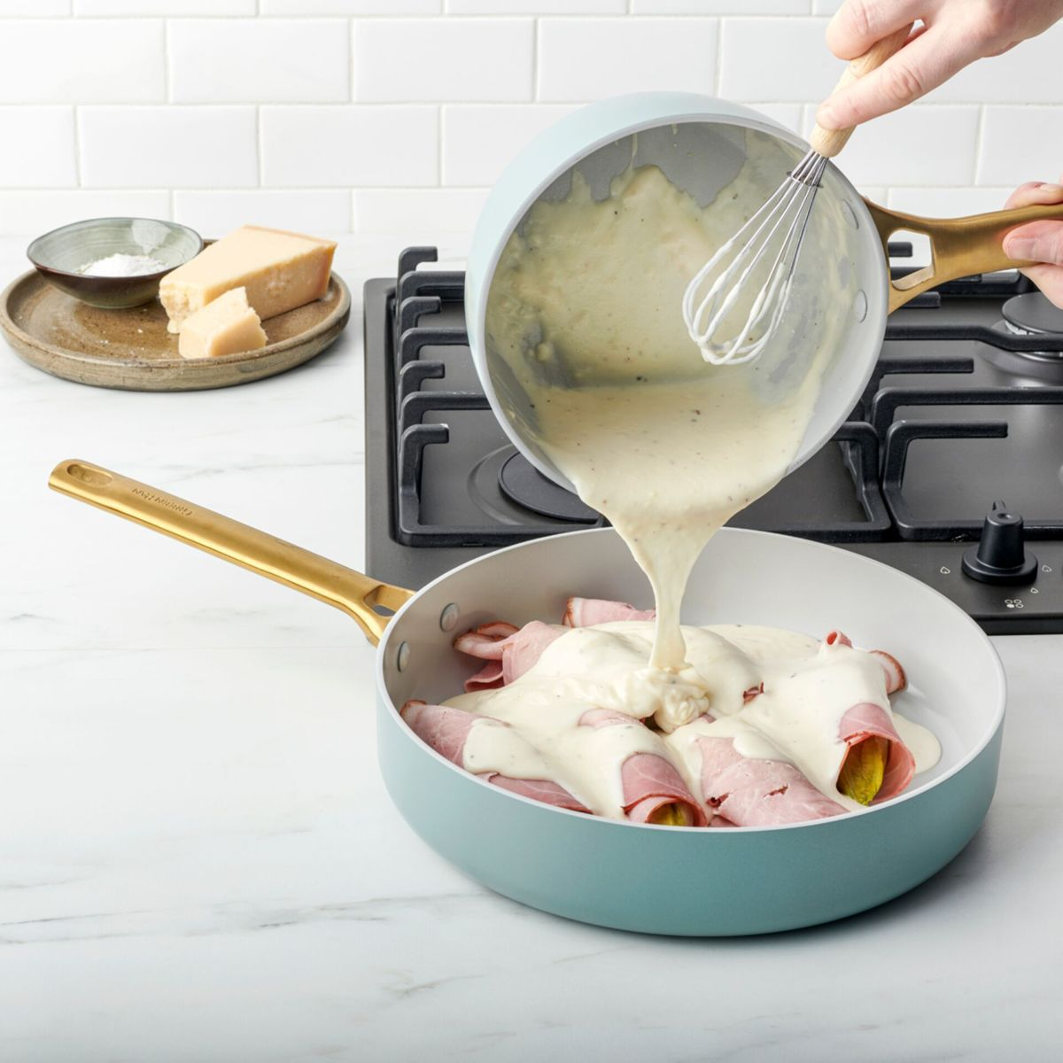 Eine Person gießt eine helle Soße aus einem Topf in eine Pfanne mit Schinkenröllchen.