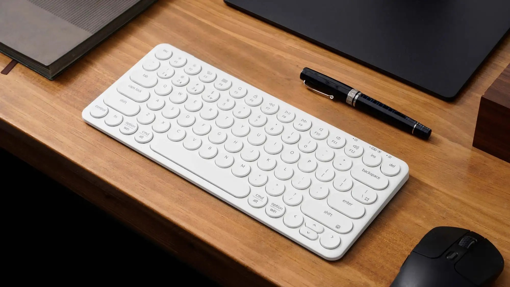 Biała klawiatura i mysz leżą na drewnianym biurku obok długopisu.