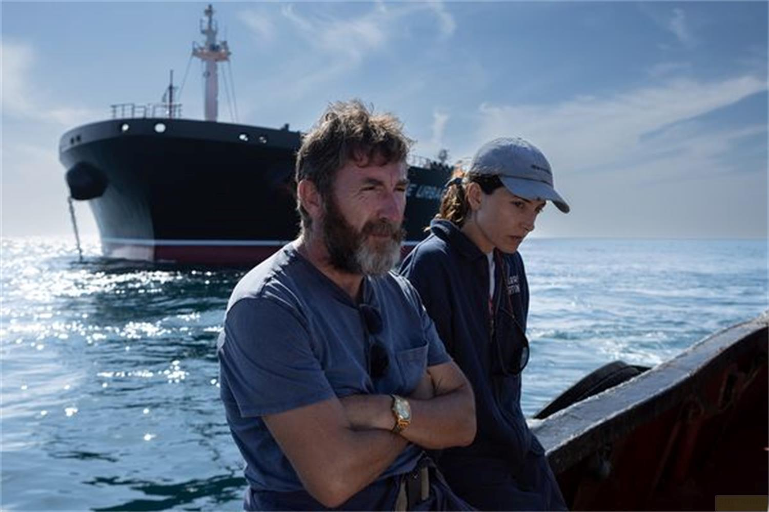 Un uomo con la barba e una donna con il berretto siedono su una barca davanti a una grande nave da carico.