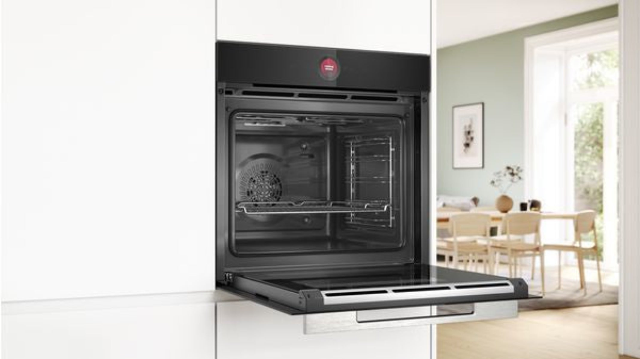 A modern built-in oven with its door open, showing a dining area in the background.