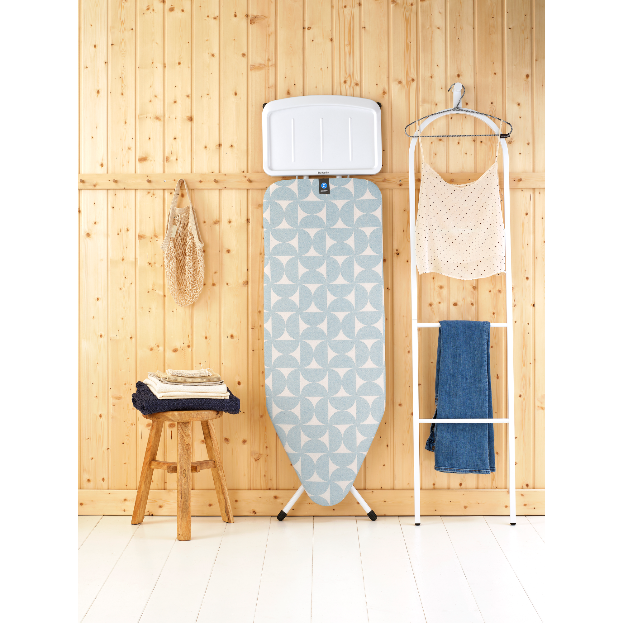 A patterned ironing board stands next to a white clothes rack with garments.