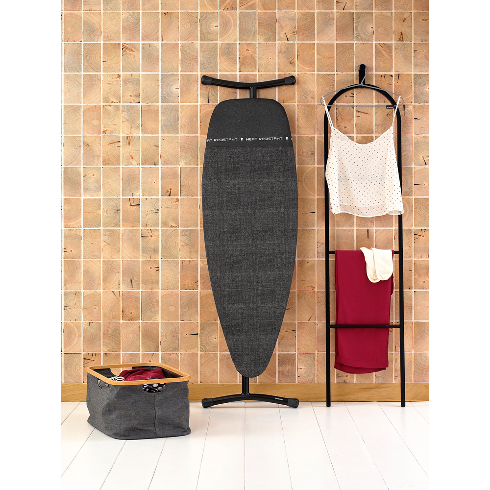 Une planche à repasser devant un mur en bois. À côté, un étendoir à linge avec des vêtements.