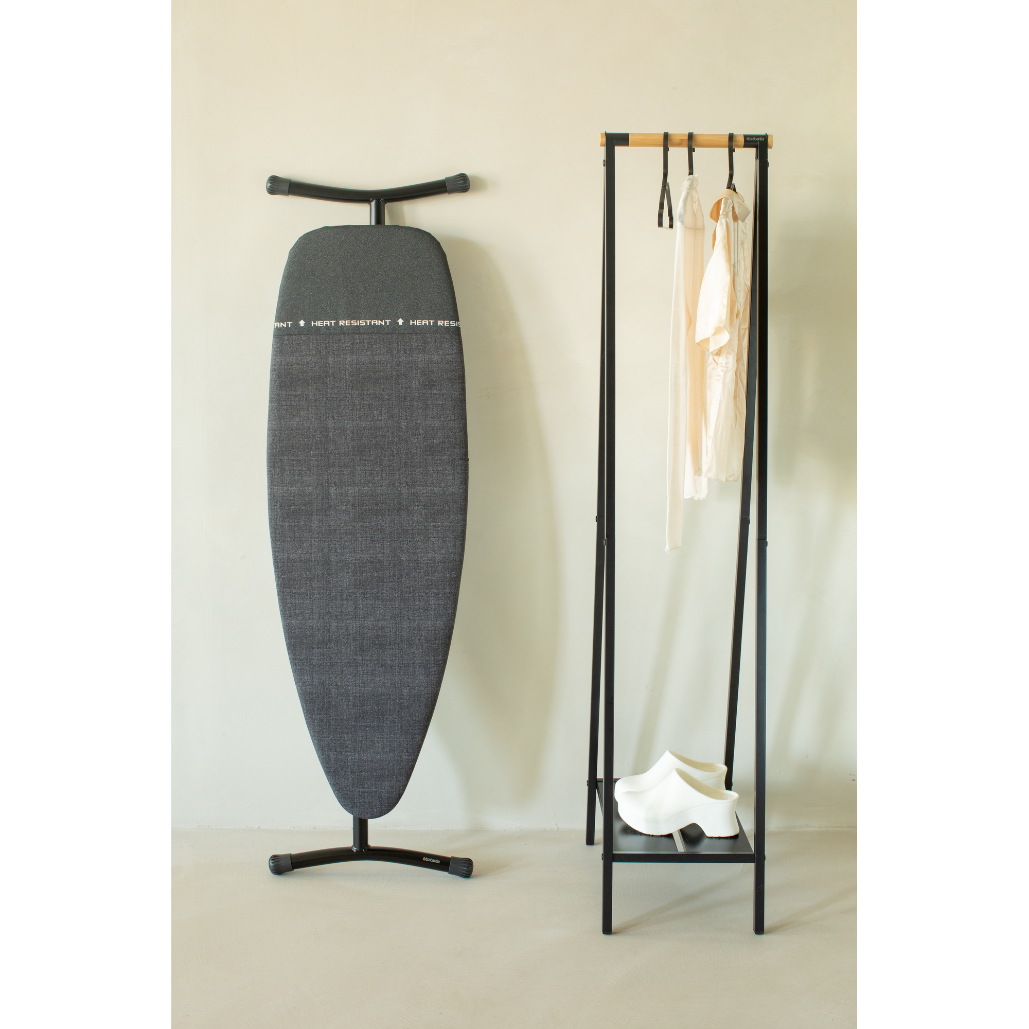 Une planche à repasser grise à côté d'un portant à vêtements avec des habits et des chaussures blanches.