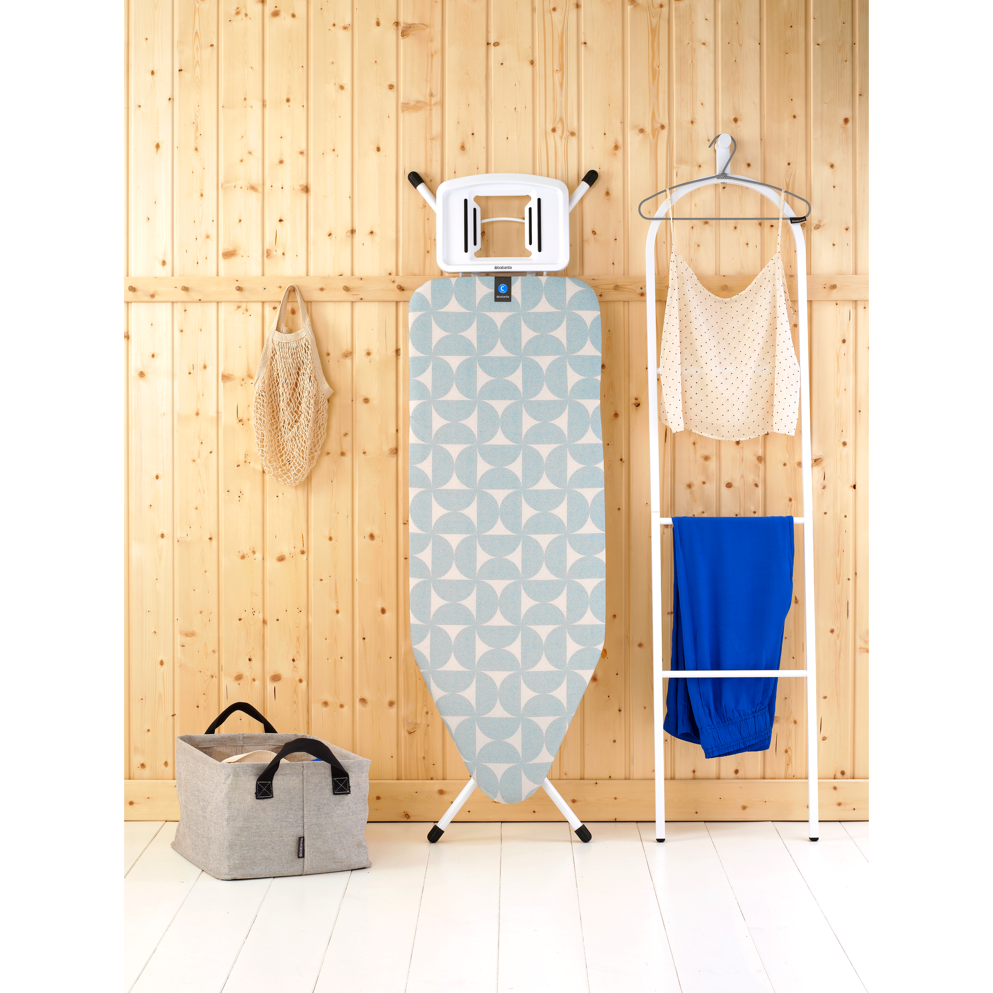 Strijkplank met patroon, wasmand, tas en kleding tegen houten wand.