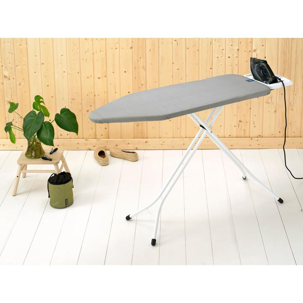Table à repasser avec housse grise, fer et plantes sur un sol en bois.