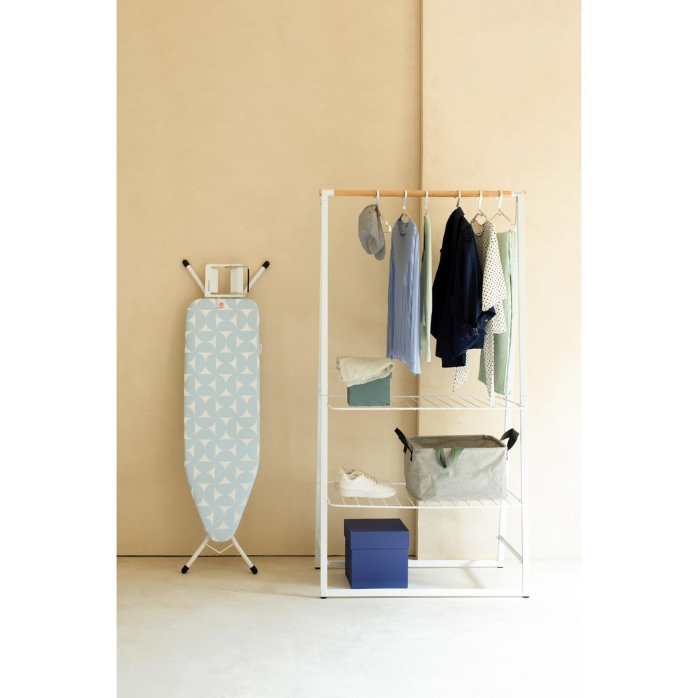 Ironing board, clothes rack with clothes, basket, shoes, boxes, wall.
