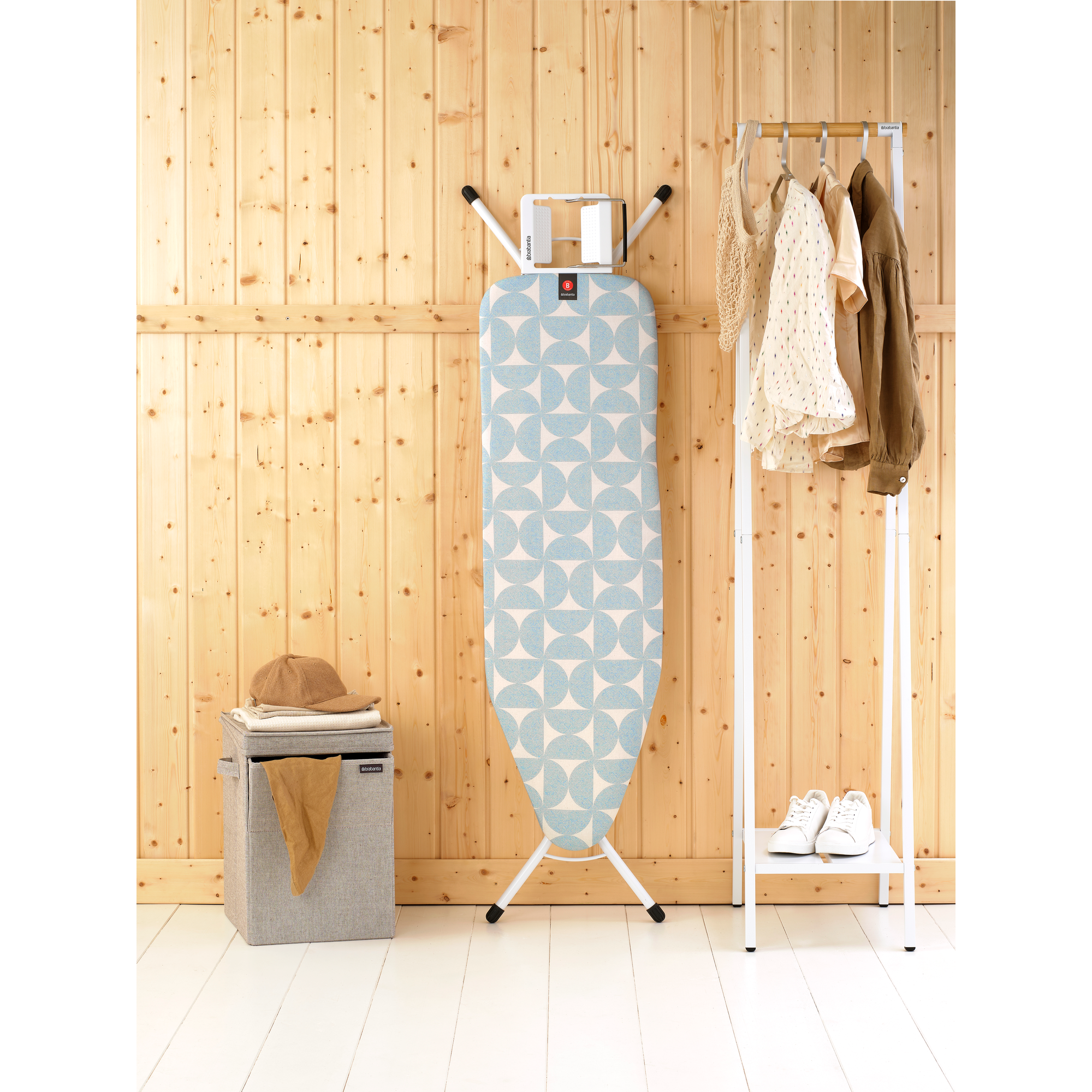 Ironing board with blue pattern, laundry basket, clothes rack with clothes and shoes.