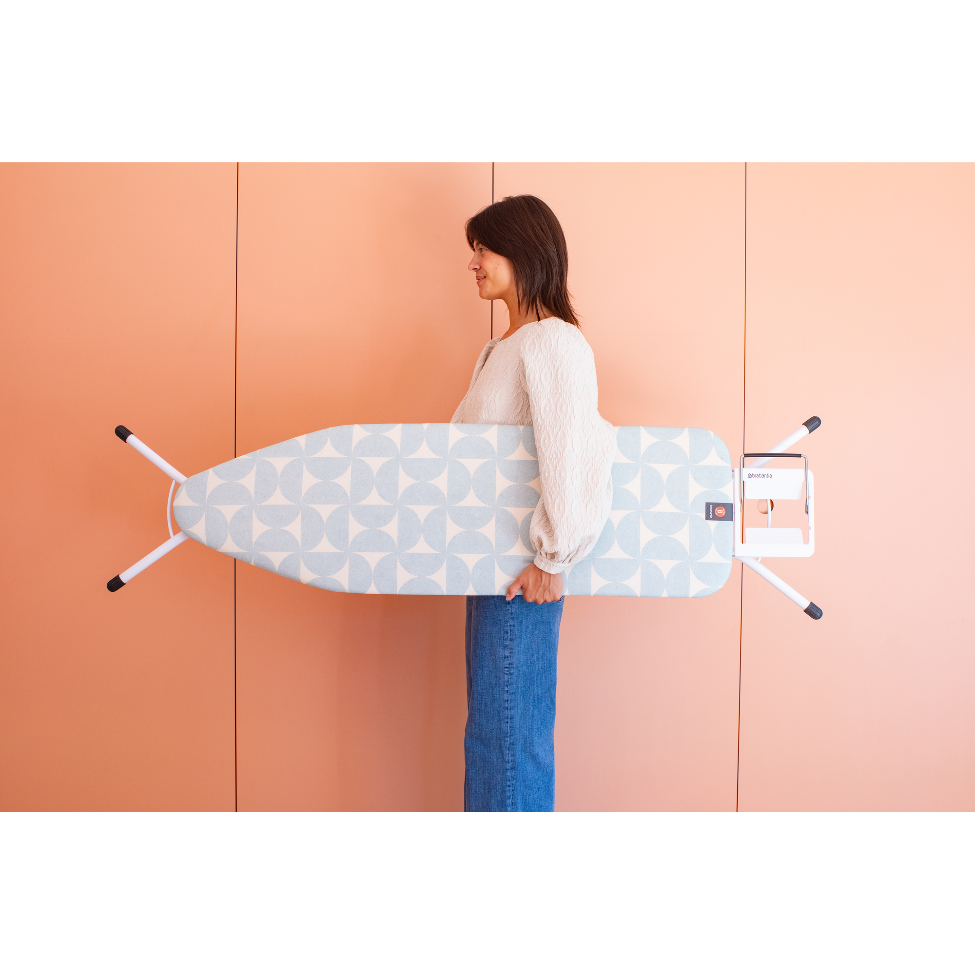 A woman holds an ironing board with a blue and white pattern against a coral wall.