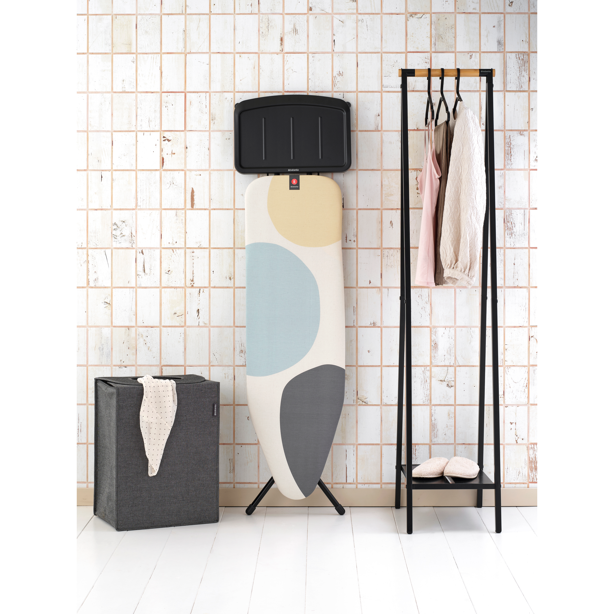Panier à linge gris, planche à repasser à motifs, portant à vêtements avec linge et chaussons.