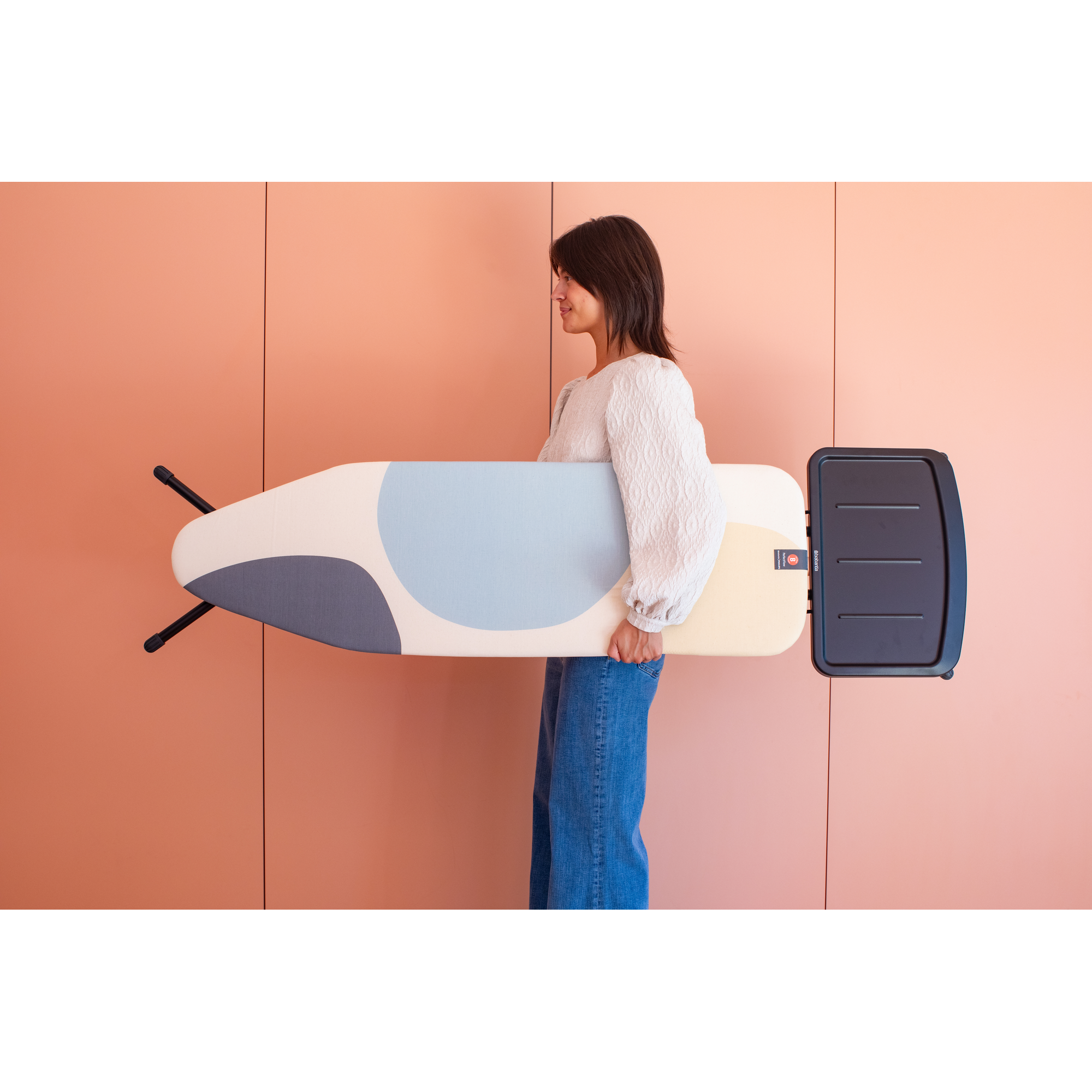 Une femme en jean et haut blanc porte une planche à repasser à motifs.