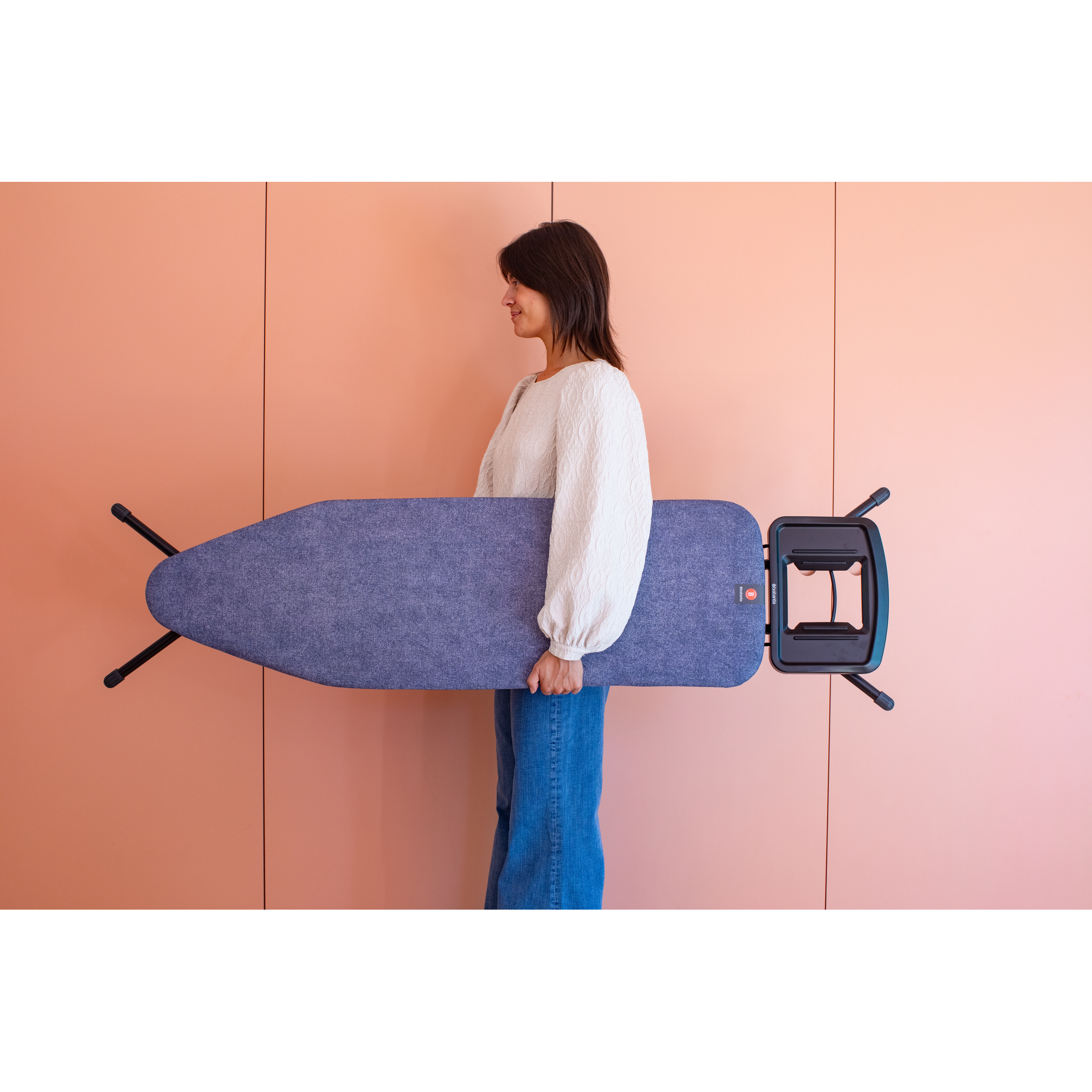 A woman holds a blue ironing board with black legs against a light pink wall.