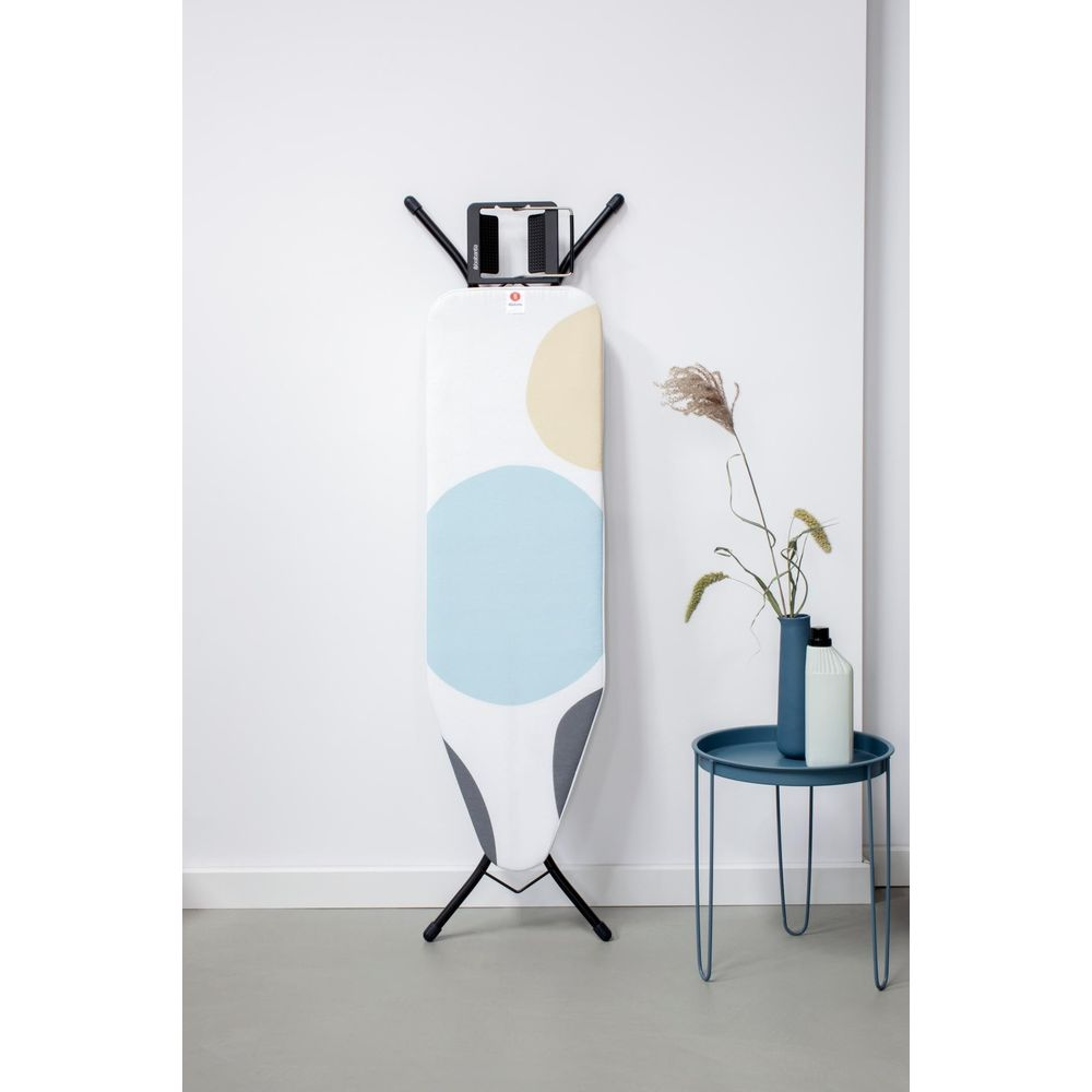 Planche à repasser avec éclaboussures de couleur contre le mur, à côté une table avec vase.