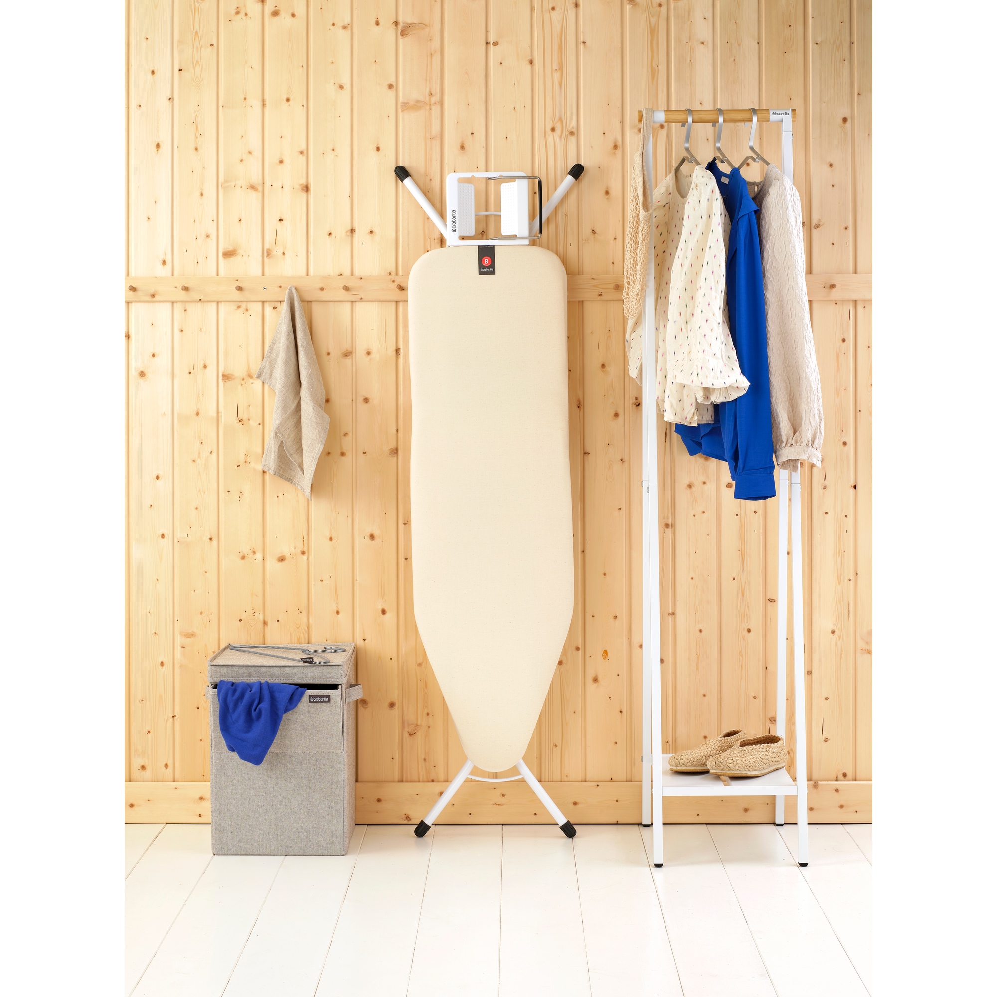 Une planche à repasser à côté d'un panier à linge et d'un portant à vêtements.