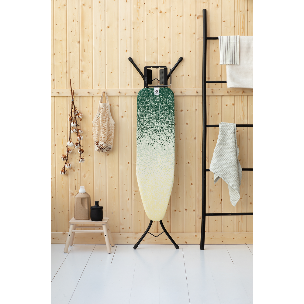 An ironing board with a green-yellow pattern stands against a wooden wall. Next to it is a black ladder rack.