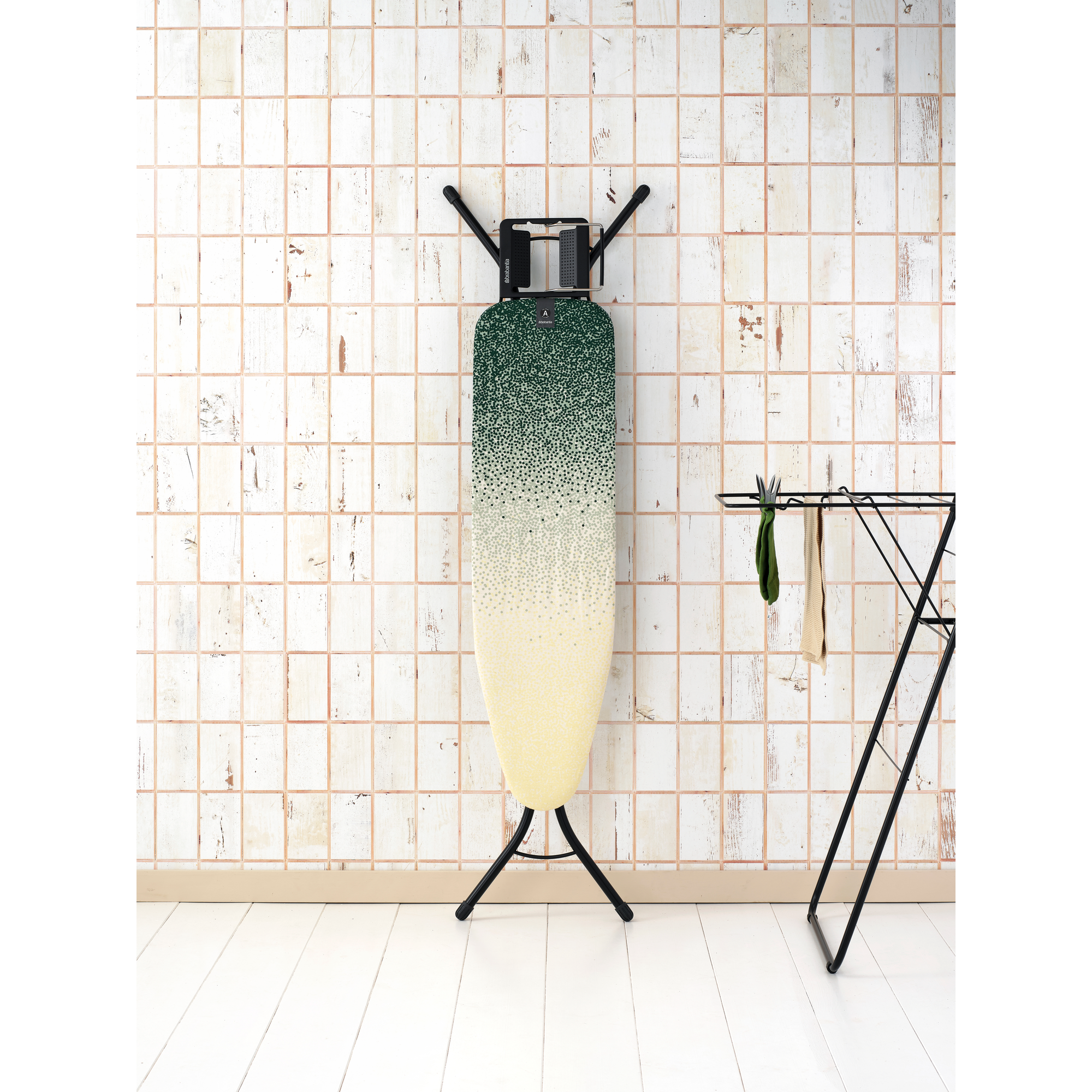 An ironing board with a green-yellow pattern stands against a tiled wall.