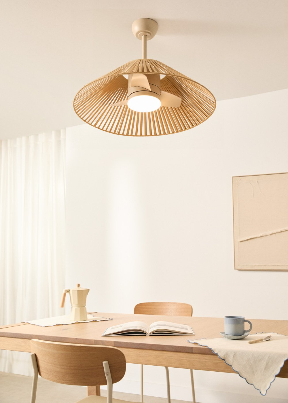Una mesa de comedor con cafetera, libro y taza bajo un ventilador de techo.