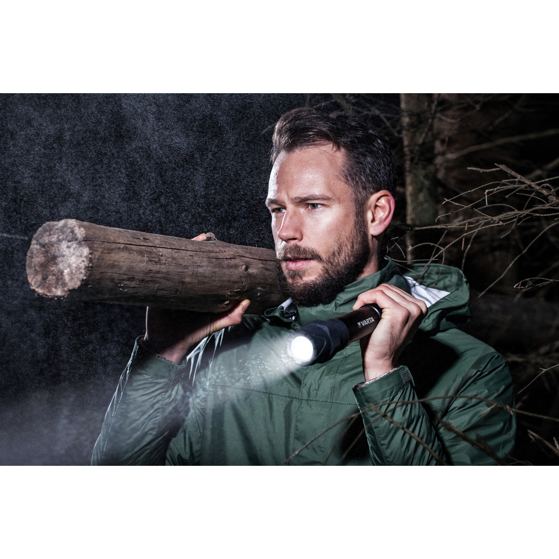 Homme en forêt portant une bûche, éclairé par une lampe torche.