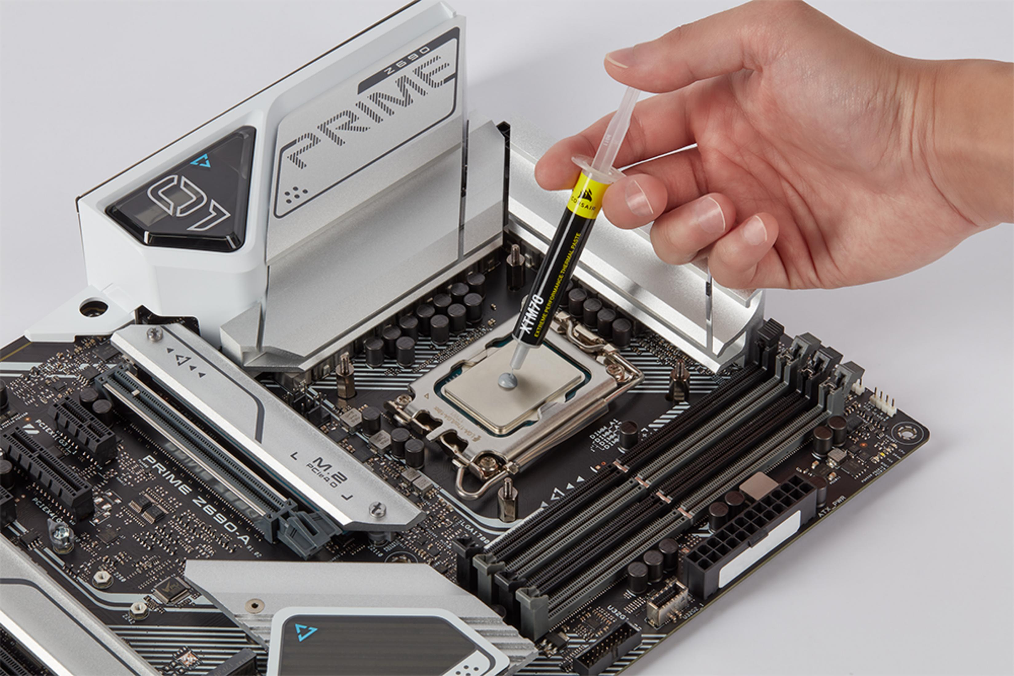 Eine Hand trägt Wärmeleitpaste aus einer Spritze auf den CPU-Sockel eines Motherboards auf.