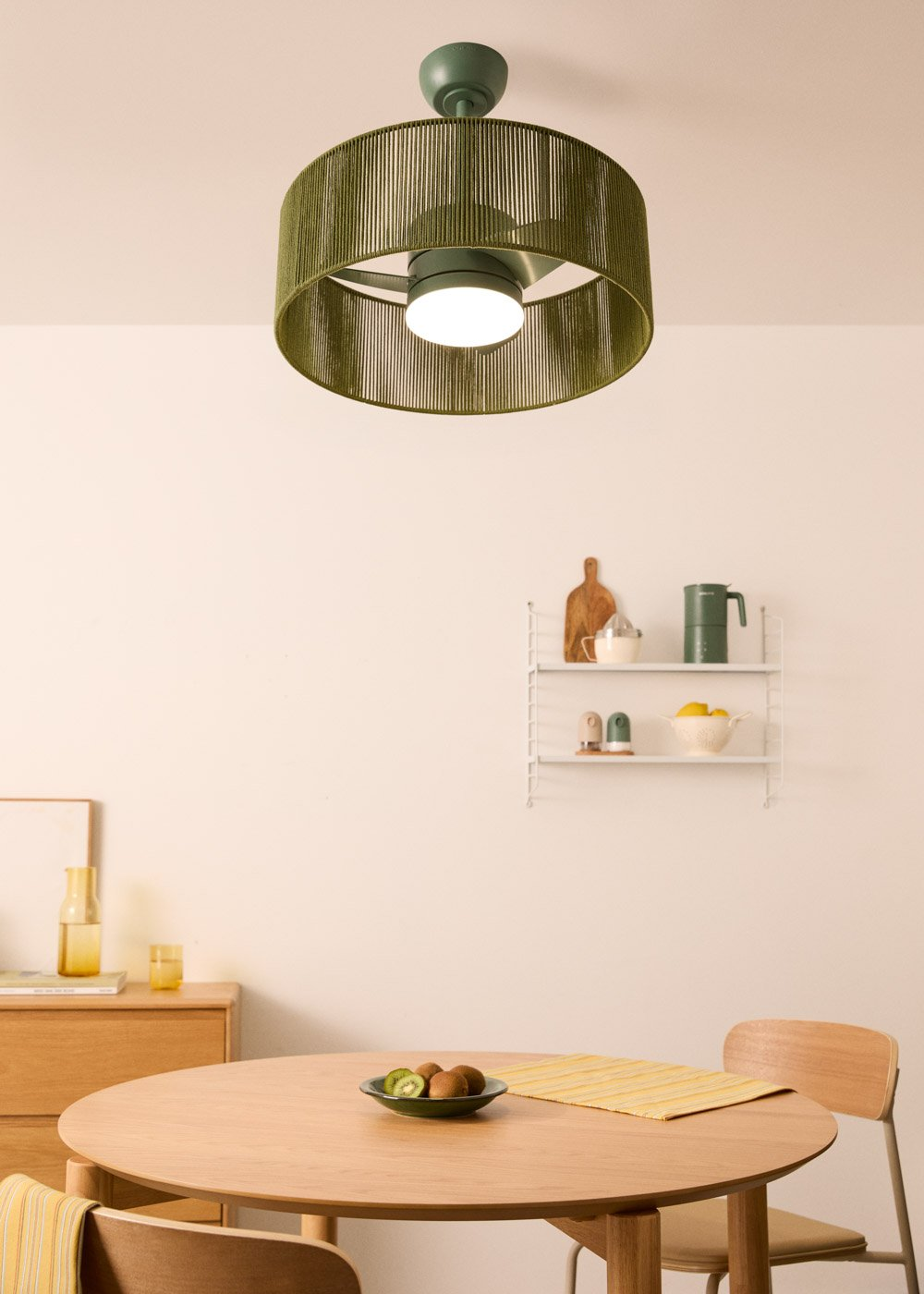 Una mesa redonda de madera con un cuenco de kiwis se sitúa ante una estantería con objetos verdes.