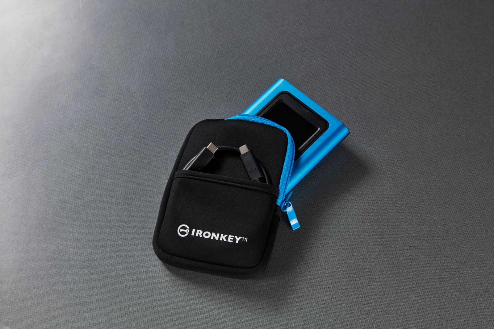 Een blauw IronKey-apparaat en de zwarte draagtas met USB-kabel liggen op een grijs oppervlak.