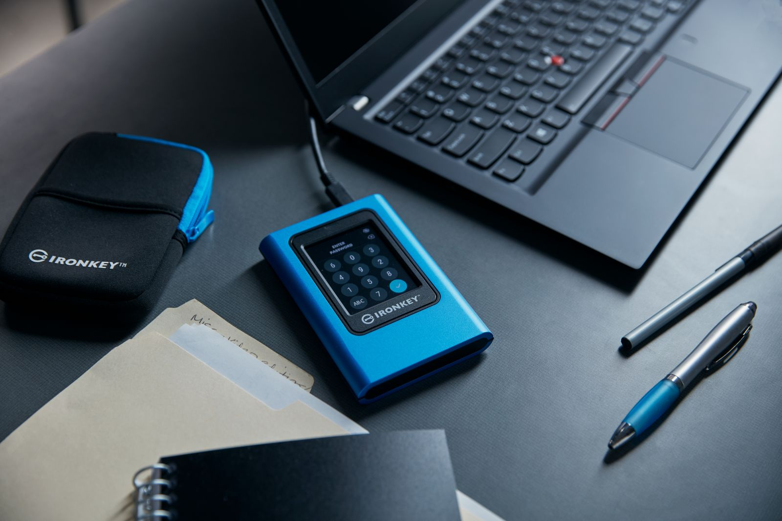 Een blauwe IRONKEY-schijf met een wachtwoordscherm, naast een laptop en pennen.