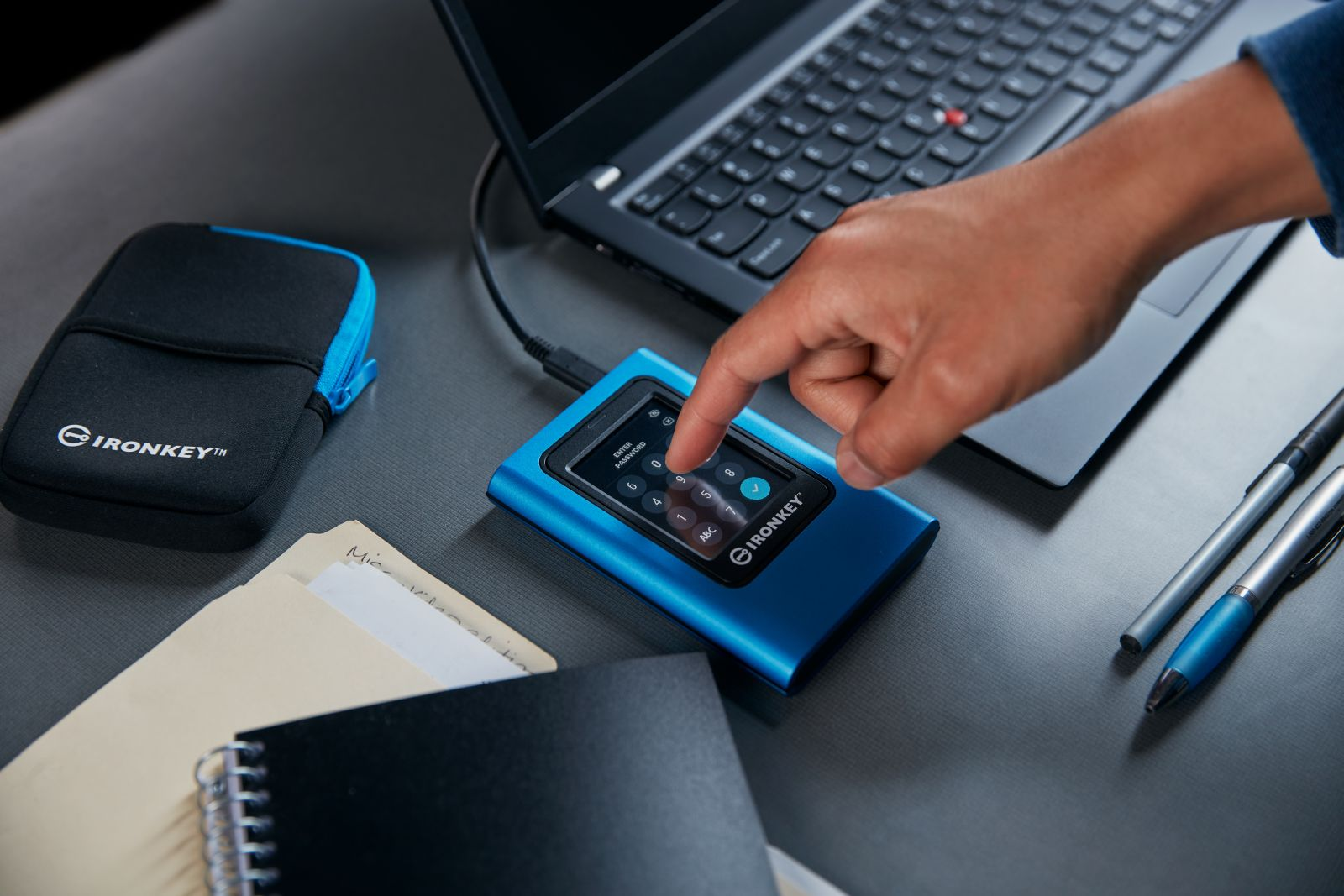 Een hand bedient een blauw extern station met wachtwoordinvoer naast een laptop.