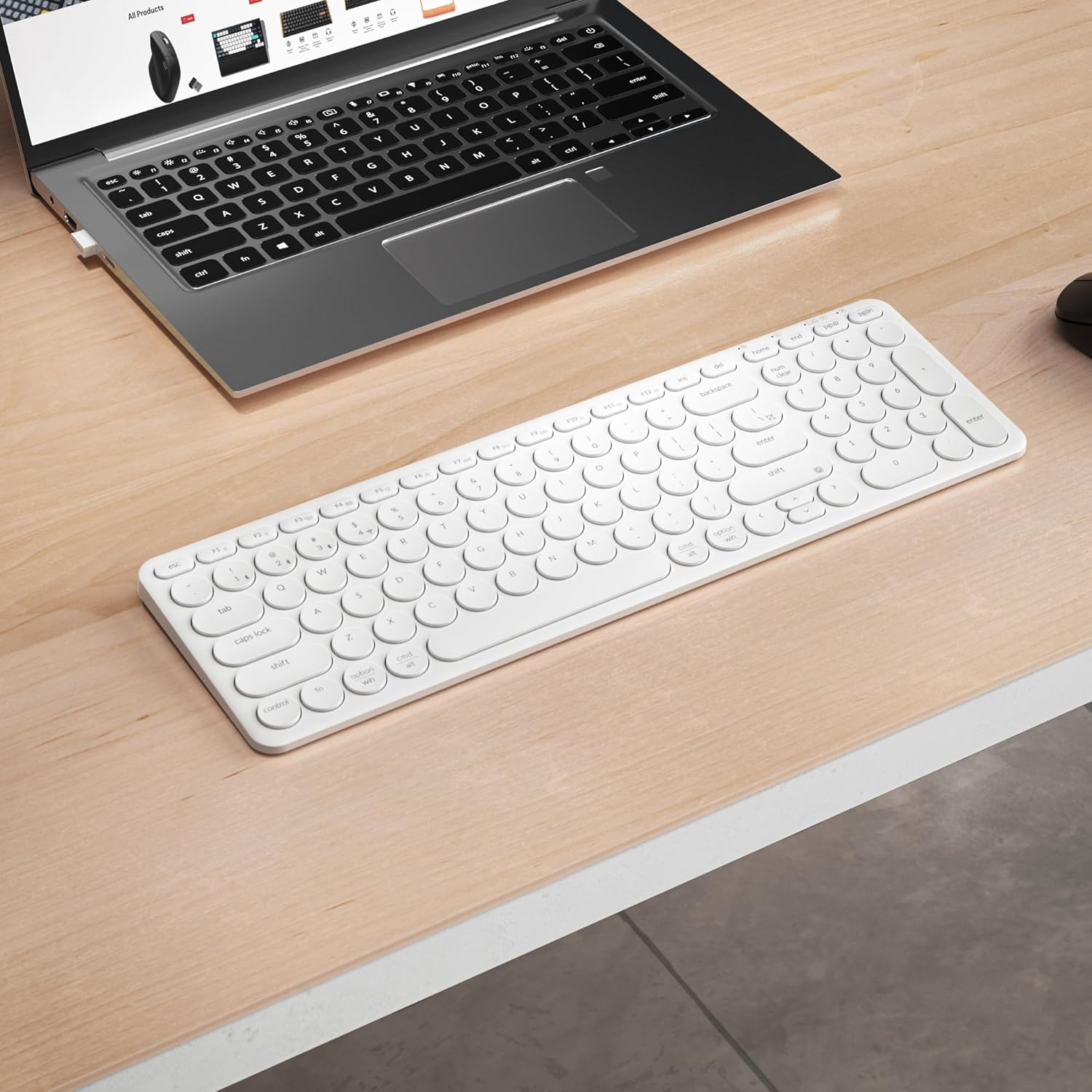 Biała klawiatura bezprzewodowa i srebrny laptop na drewnianym biurku. Widoczna jest czarna mysz.