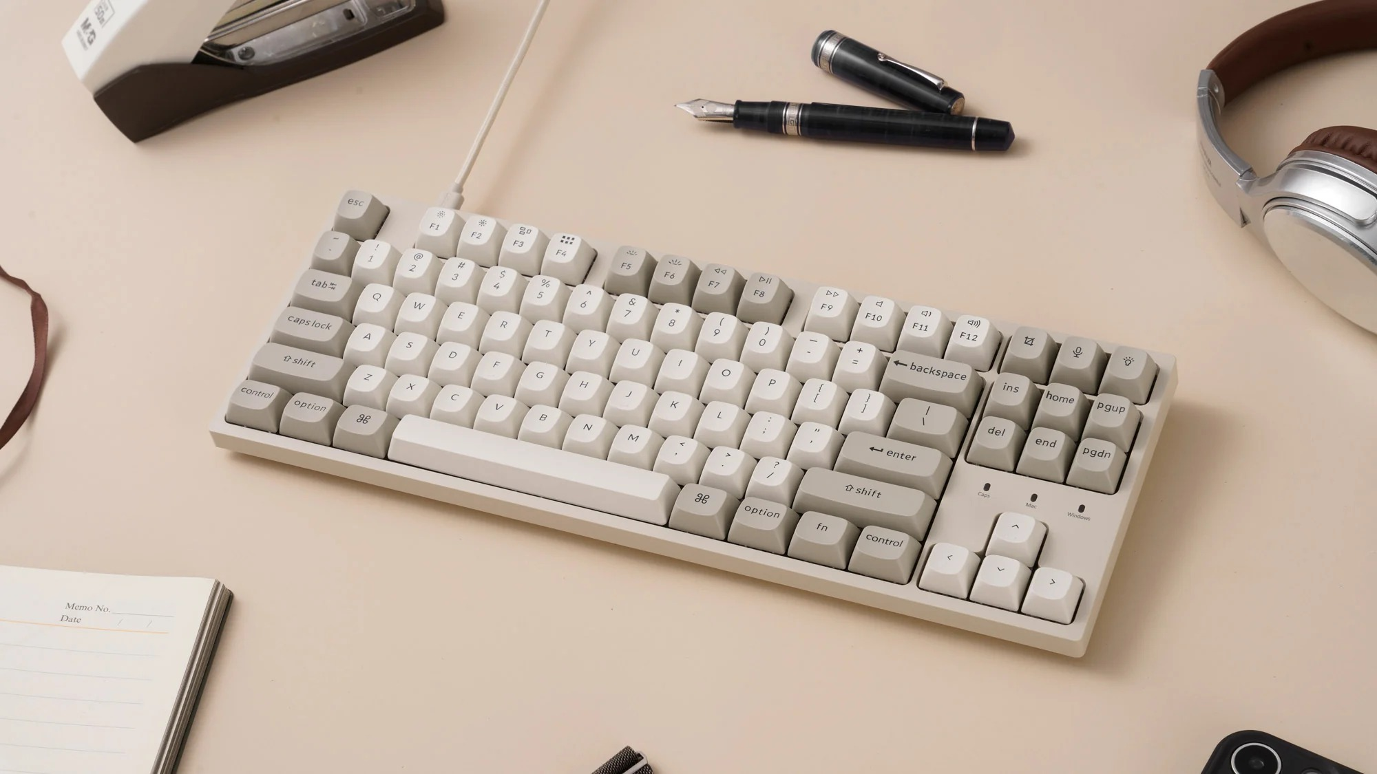 Biała klawiatura z długopisem, słuchawkami, notatnikiem i zszywaczem na beżowej powierzchni.