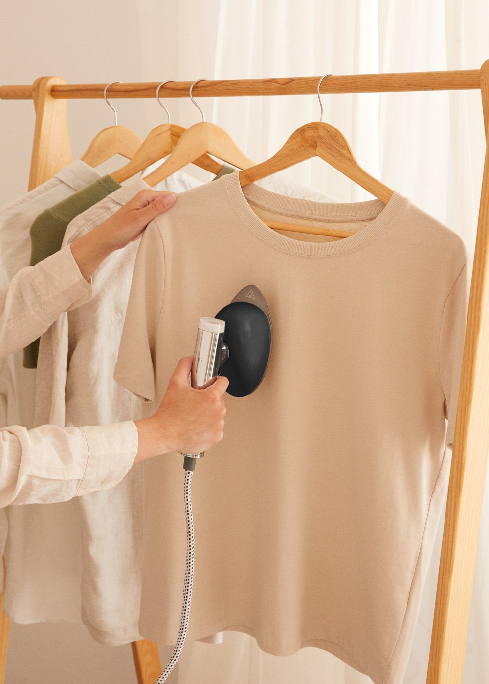 Una mano usa un vaporizador de mano en una camiseta beige colgada en una percha.