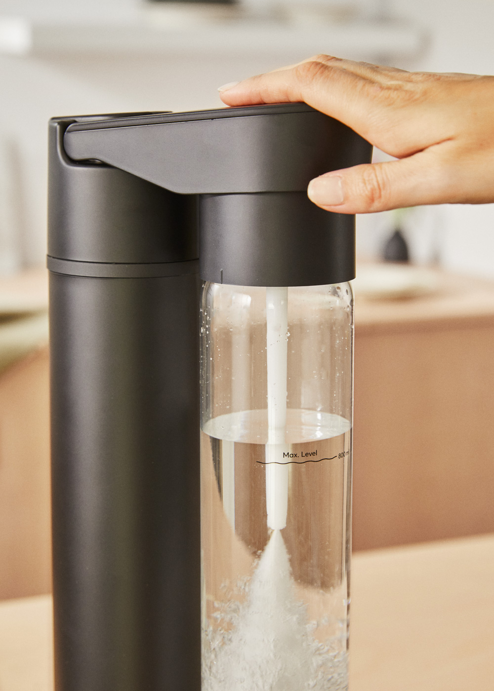 Una mano presiona un carbonatador de agua negro con recipiente de vidrio.