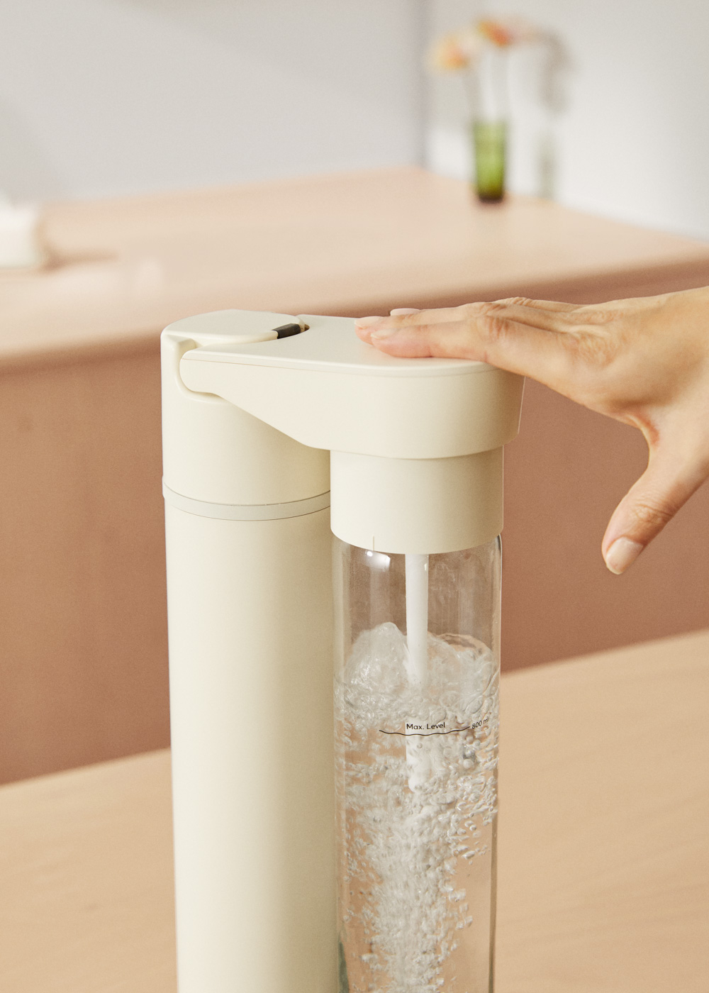 Una mano presiona una máquina de soda color crema con una botella de vidrio llena de burbujas.