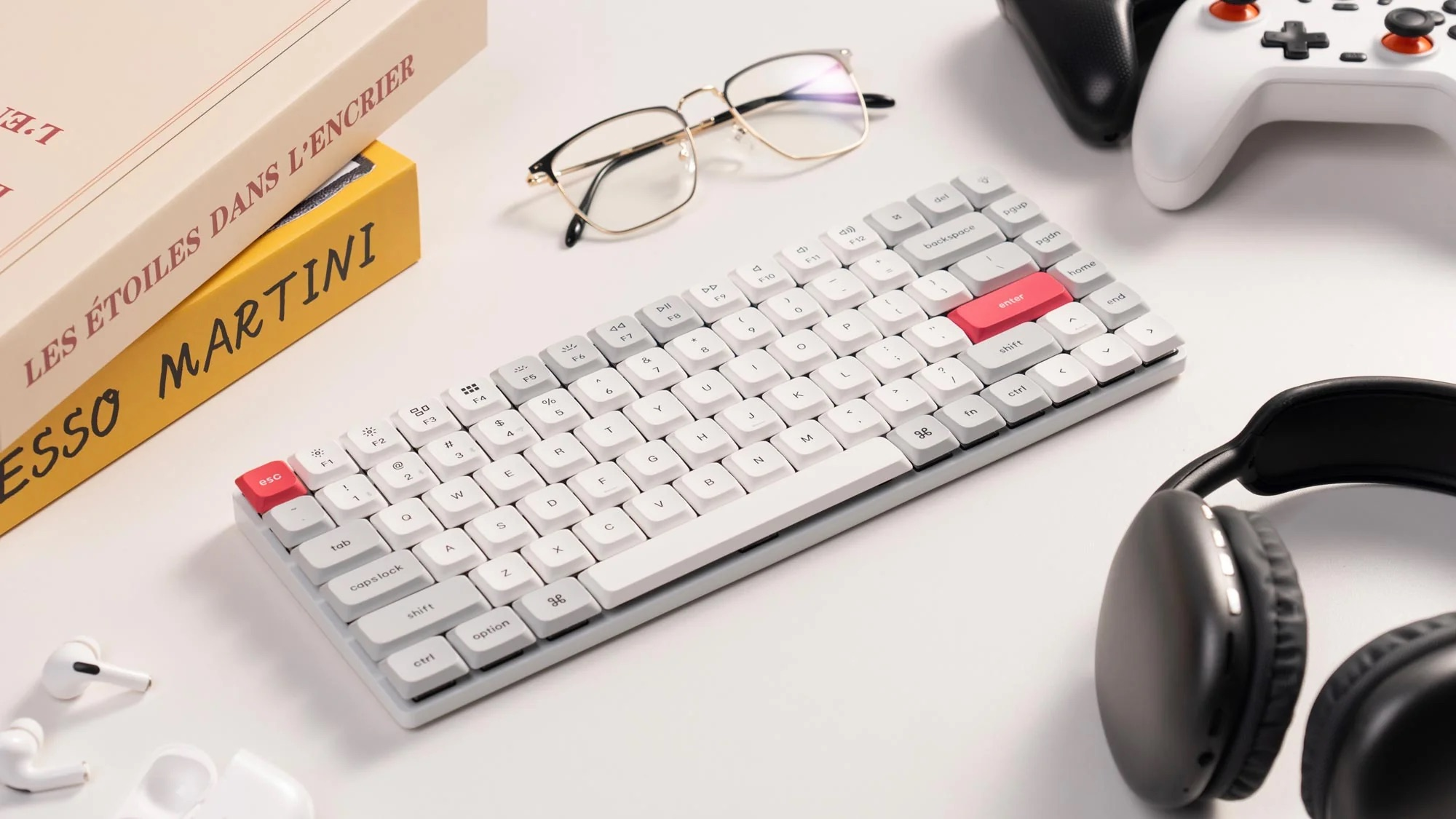 Biała klawiatura z czerwonymi klawiszami Enter/Esc, okularami, słuchawkami, kontrolerami i książkami.