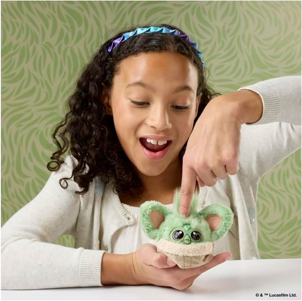 Niña con diadema sostiene una pequeña criatura verde con ojos grandes.