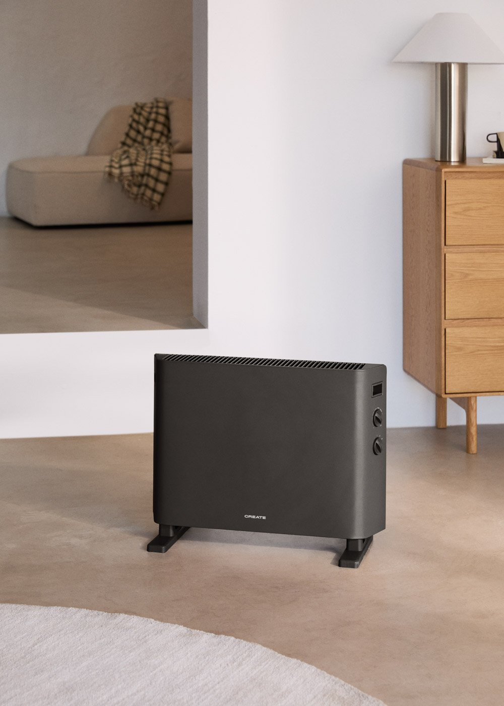 Radiador oscuro con perillas de control en el suelo, frente a mueble de madera con lámpara.
