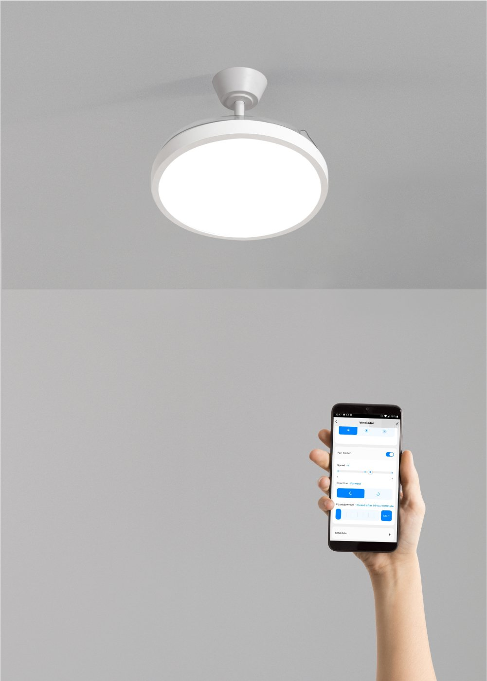 Un ventilador de techo blanco con luz está montado en un techo gris. Una mano sostiene un teléfono inteligente mostrando controles del ventilador.