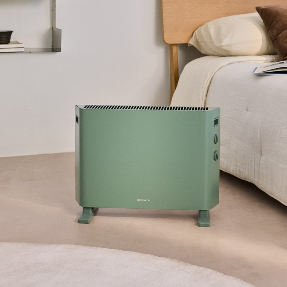Un calefactor verde menta sobre una alfombra clara en un dormitorio. Una cama con ropa de cama blanca y cabecero de madera al fondo.