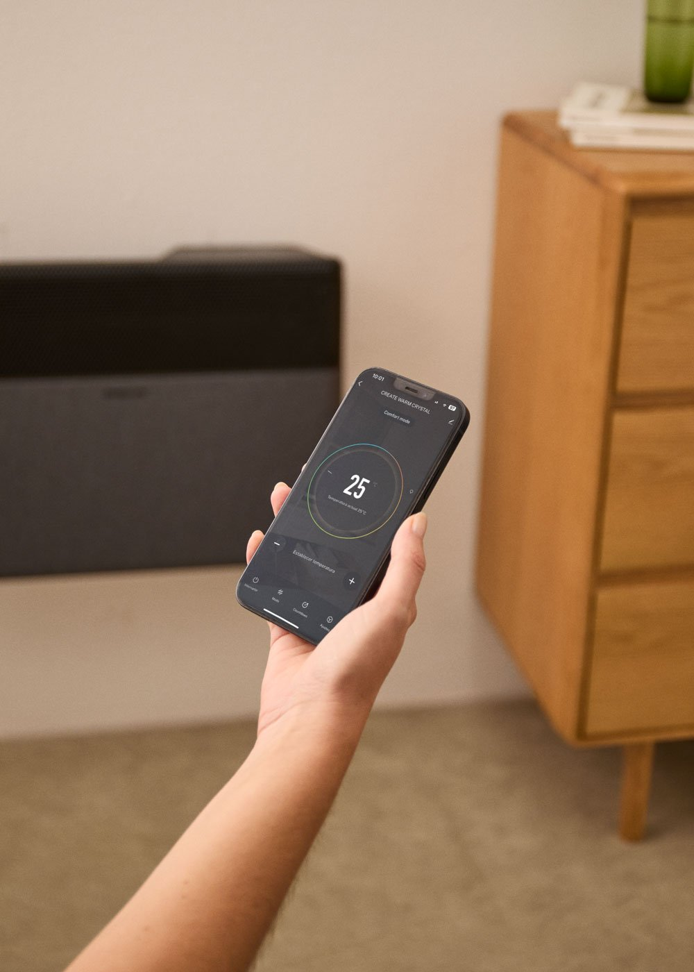 Mano sostiene smartphone con control de calefacción, muestra 25°C. Radiador al fondo.