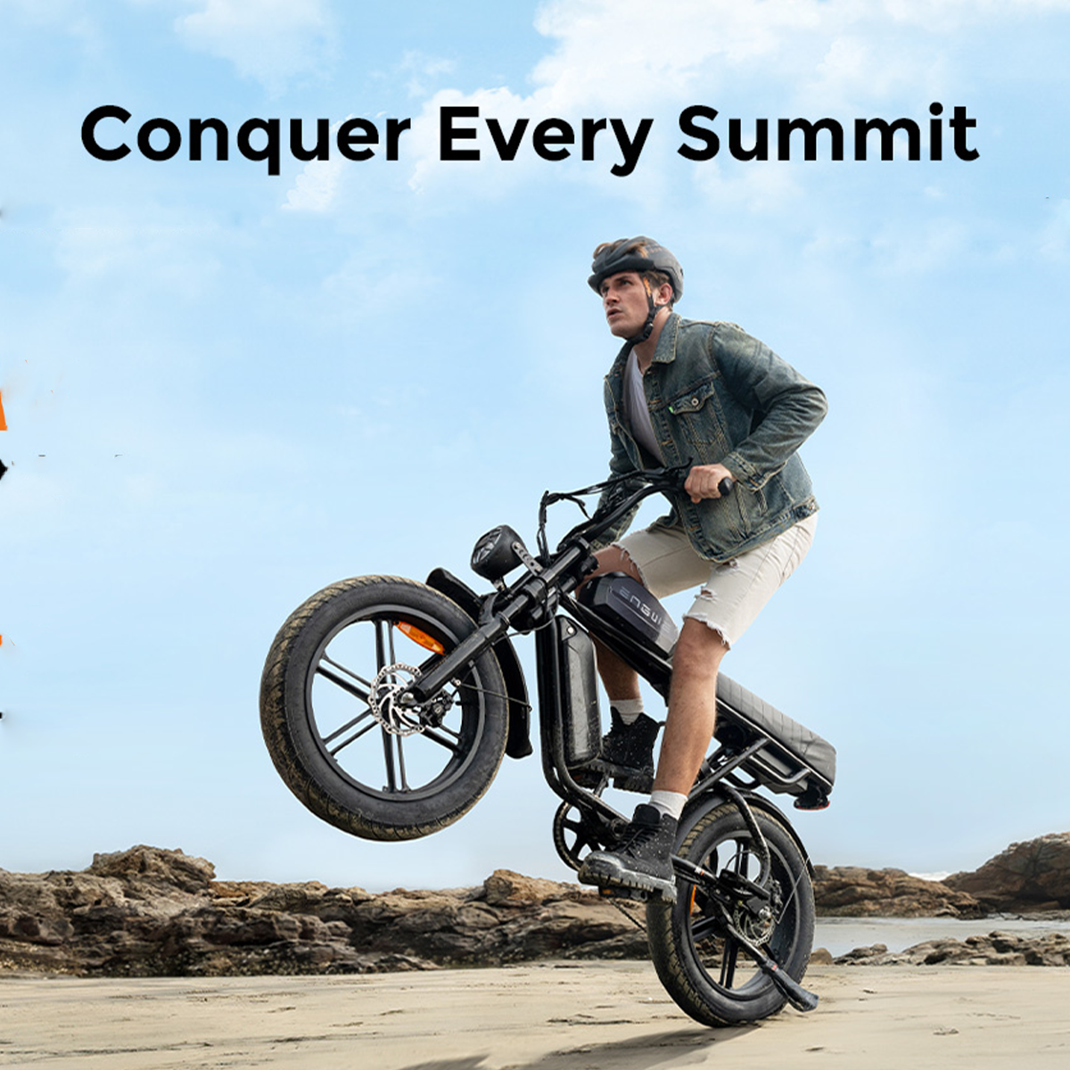 Mężczyzna na rowerze elektrycznym robi wheelie na piaszczystej plaży z kamienistym tłem.