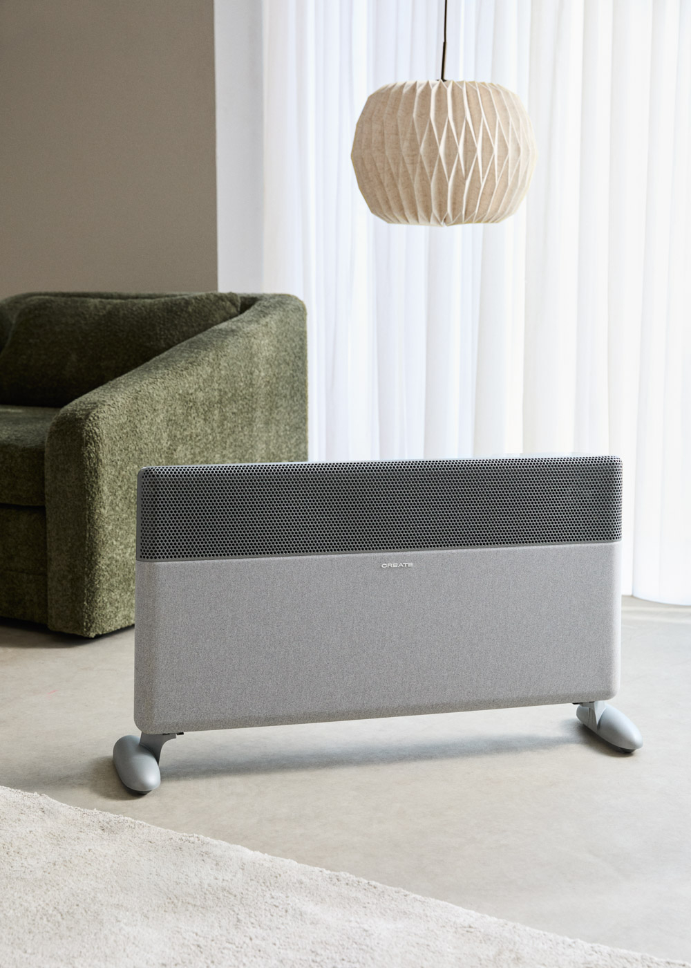 Un radiador gris con ruedas se sitúa ante un sillón verde y cortinas transparentes.