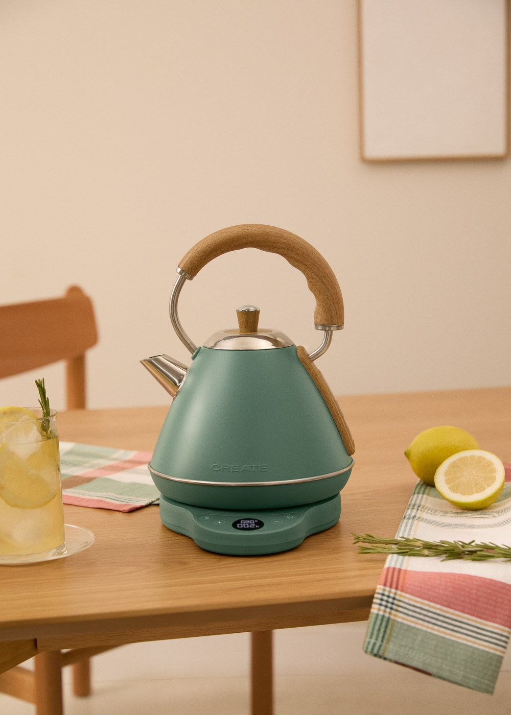 Un hervidor verde con asa de madera está sobre una mesa junto a un vaso de agua y limones.