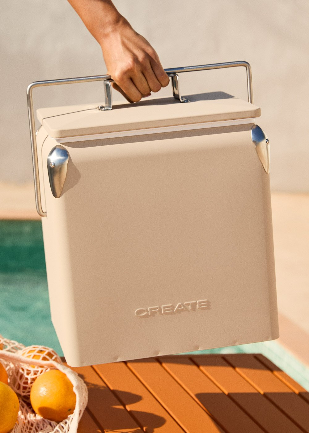 Una mano sostiene una hielera beige con asas cromadas, junto a una piscina.