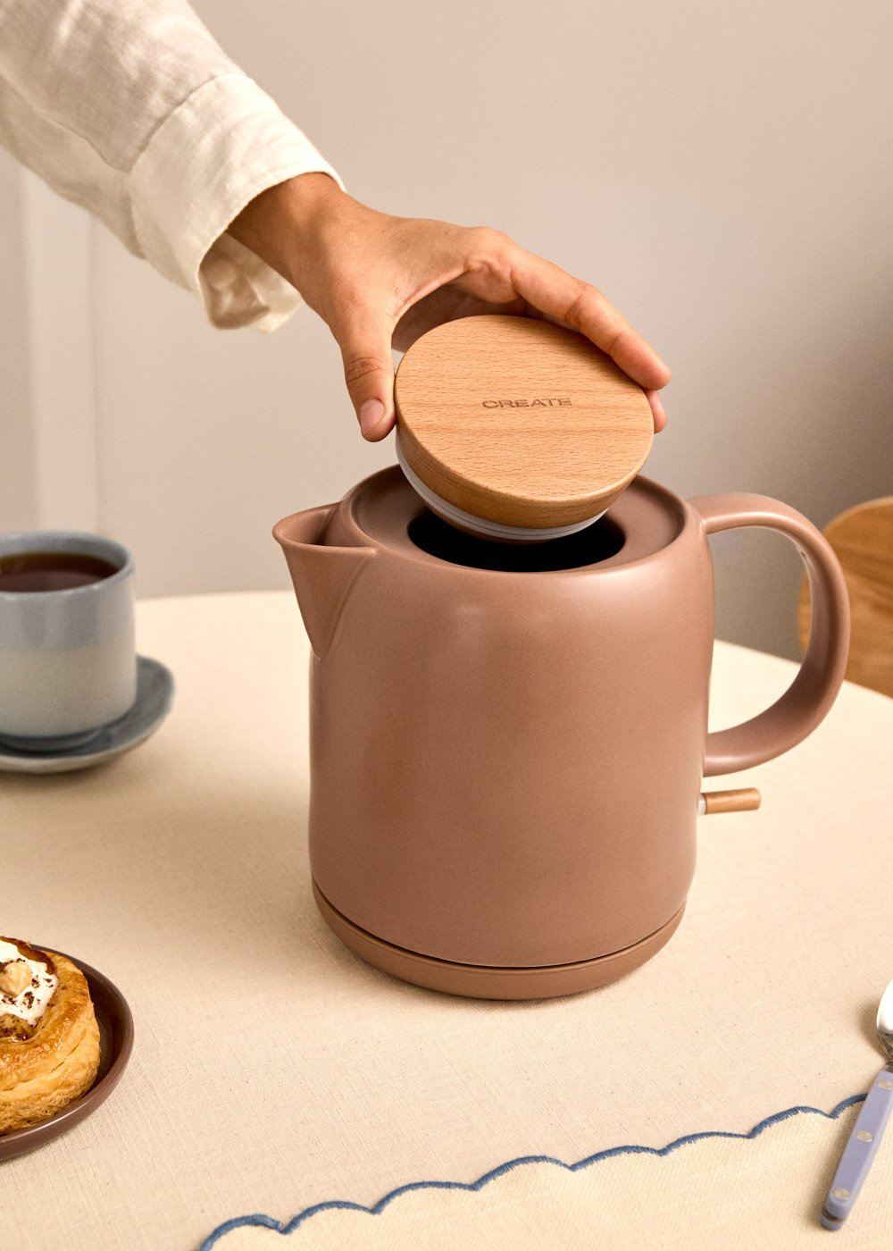 Una mano coloca una tapa de madera sobre un hervidor marrón junto a una taza de té.