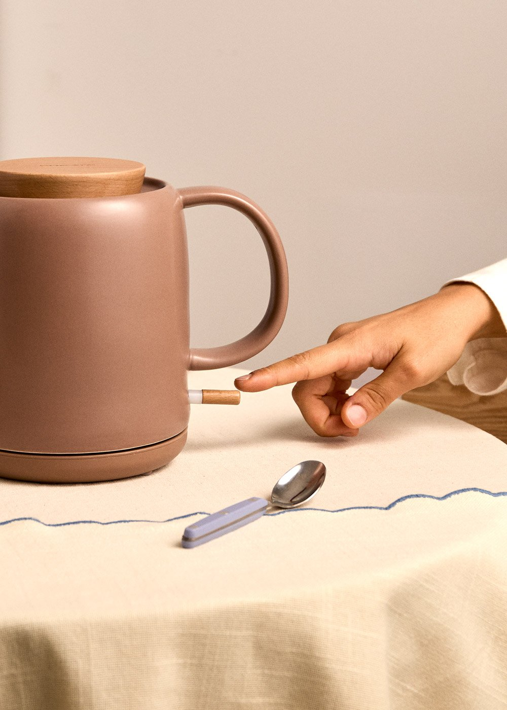 Una mano presiona el interruptor de una tetera color topo con tapa de madera.