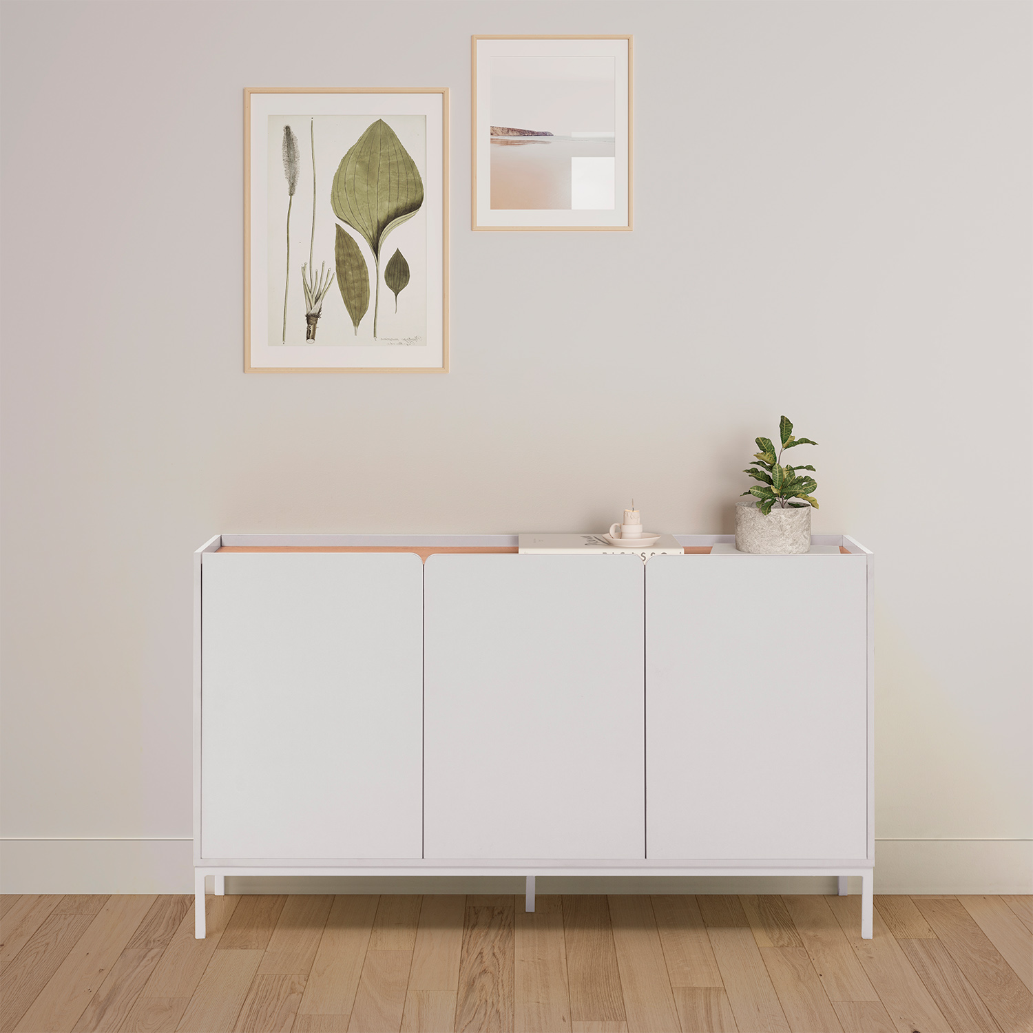Un aparador blanco de tres puertas sobre suelo de madera, con cuadros y una planta encima.