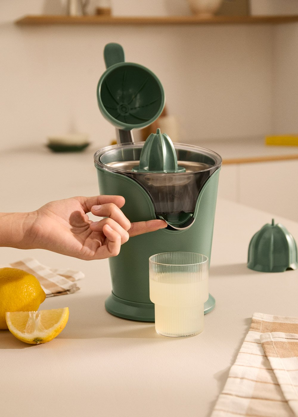 Exprimidor verde con limones, vaso de jugo. Mano presiona botón.