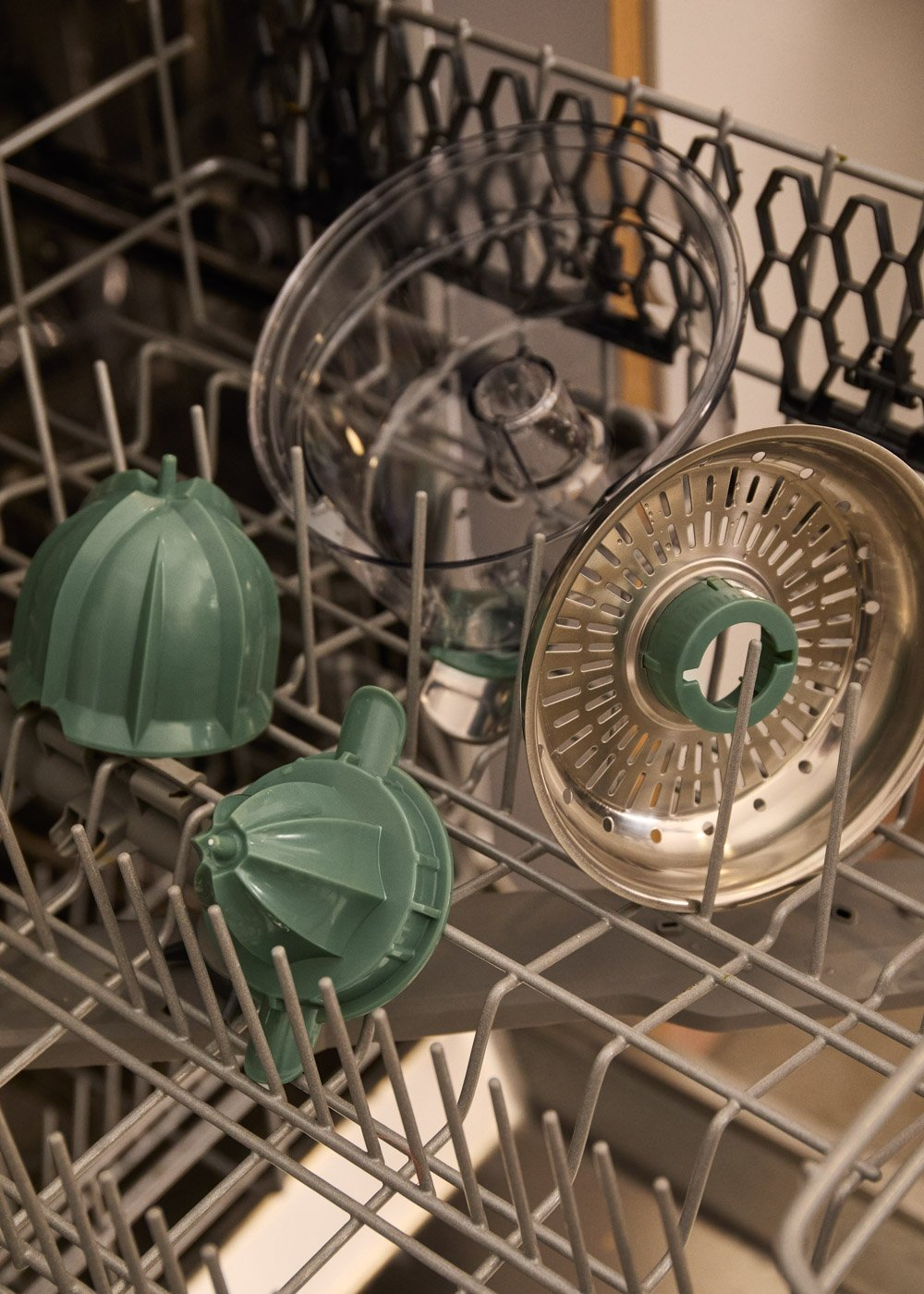Partes verdes de procesador de alimentos y colador metálico en cesta de lavavajillas.