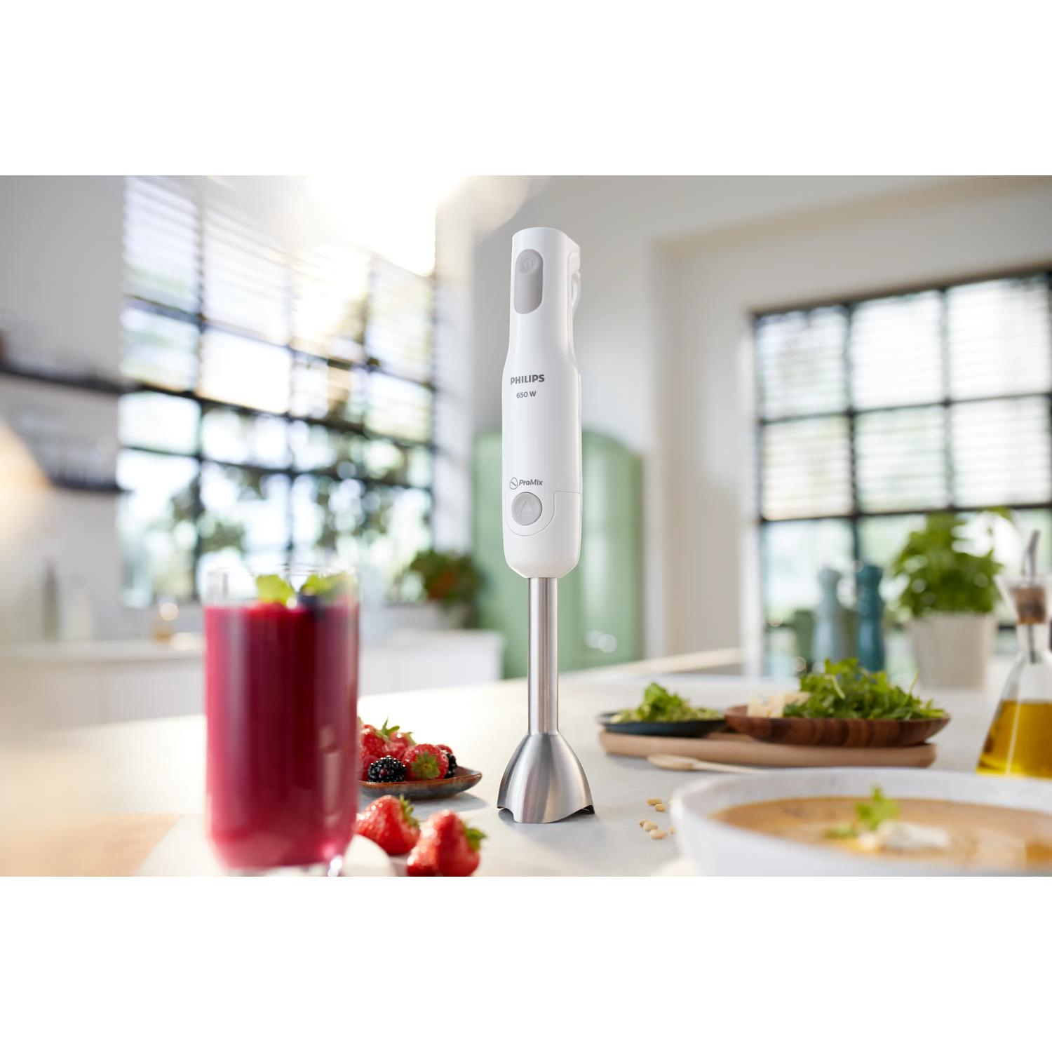 Un frullatore a immersione bianco poggia sul bancone della cucina accanto a un bicchiere di succo e bacche.