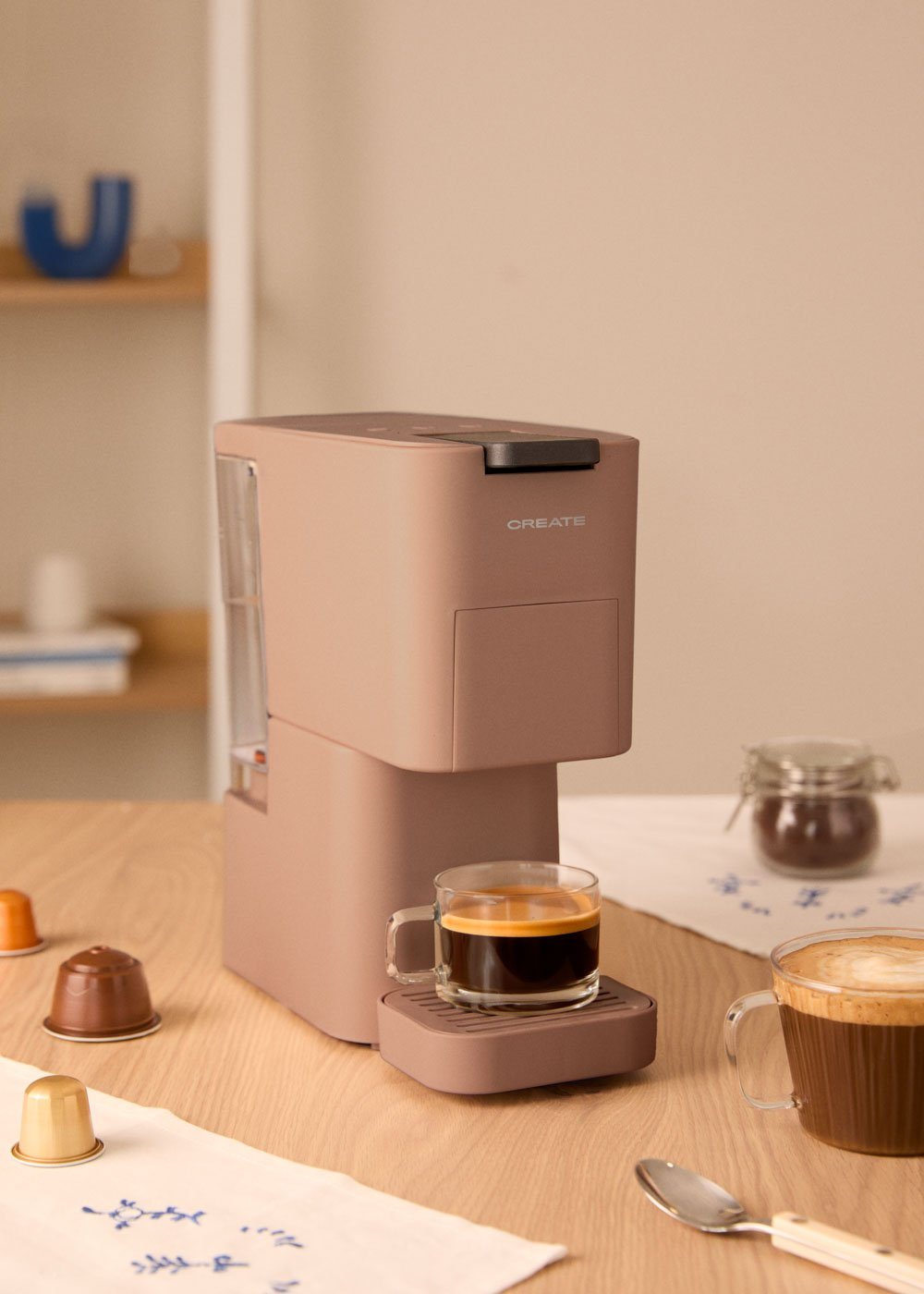 Una cafetera rosa con una taza de espresso, cápsulas de café y una taza de café sobre una mesa.