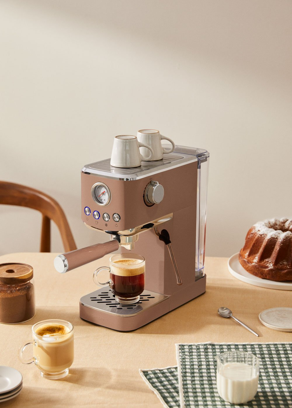 Una máquina de espresso color oro rosa con dos tazas encima, café recién hecho y pastel.